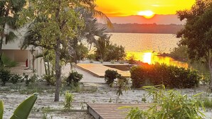 Eine Person sitzt auf einer Holzterrasse mit einem schwarzen Hund und blickt auf einen Strand mit Palmen und einem Sonnenuntergang.