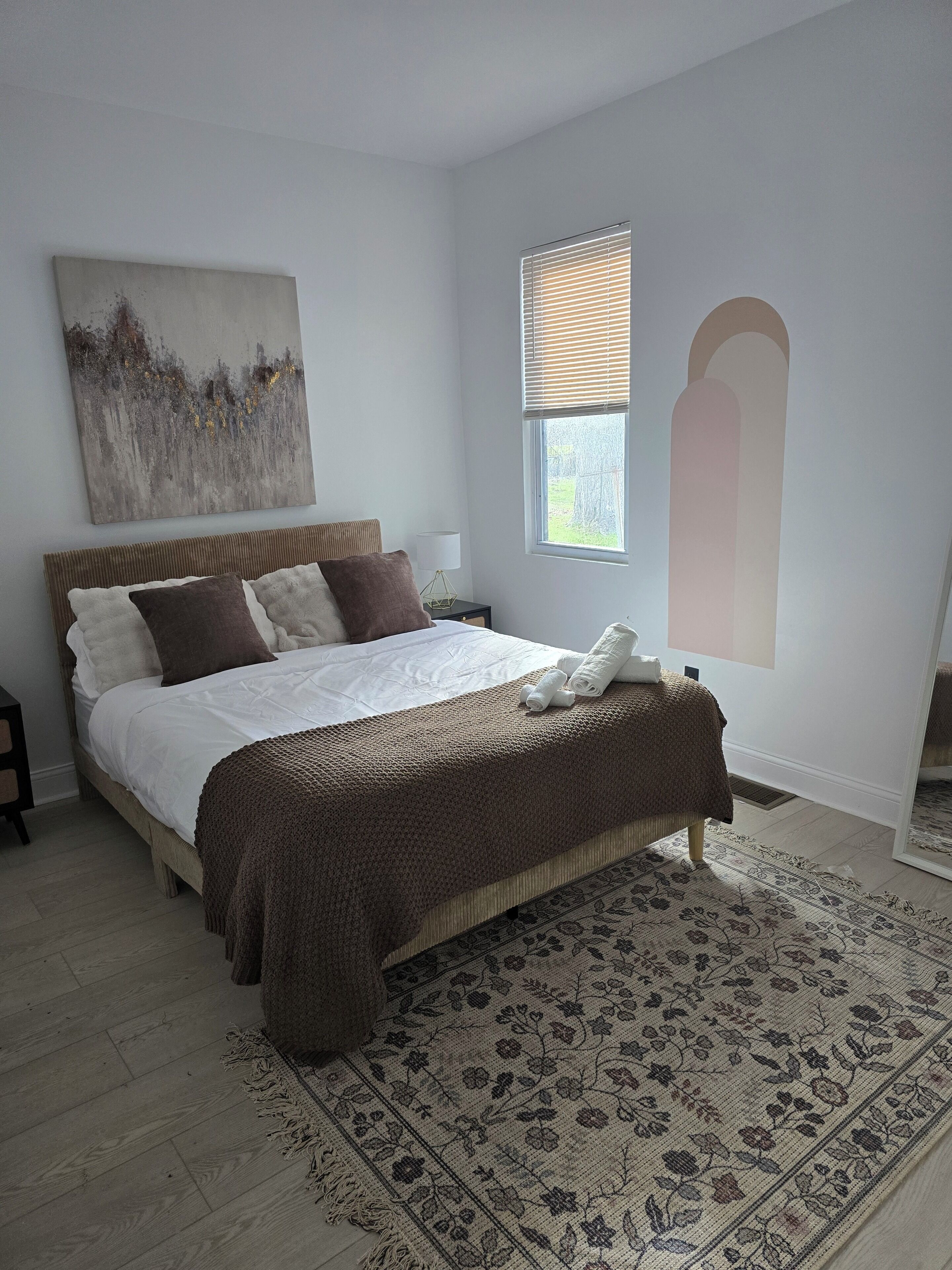 Une chambre à coucher avec un grand lit, une table de chevet, une fenêtre avec des stores, une œuvre d’art accrochée au mur et un tapis à motifs.