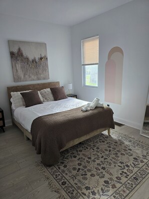 A bedroom with a large bed, a nightstand, a window with blinds, a wall-mounted artwork, and a patterned rug.