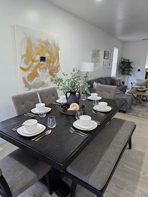A dining area with a dark wooden table set for four, surrounded by grey upholstered chairs. A large abstract painting adorns the wall behind the table.