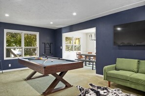 A pool table in a room with dark walls, a green sofa, and a flat-screen TV mounted on the wall.