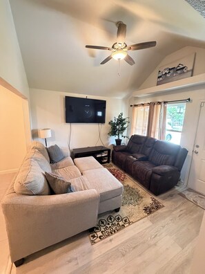 A living room with a sofa, a recliner, a flat-screen TV, a ceiling fan, and a window with curtains. - 3BR Near Arlington World Cup & Downtown FTW (Fort Worth)