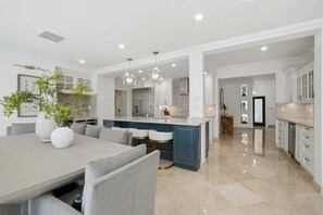 Una cocina moderna con armarios blancos, una isla central, iluminación colgante y una zona de comedor con mesa y sillas.