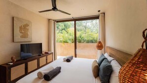 Ein Schlafzimmer mit Bett, Fernseher, Deckenventilator und einem Balkon mit Blick auf Grünflächen.