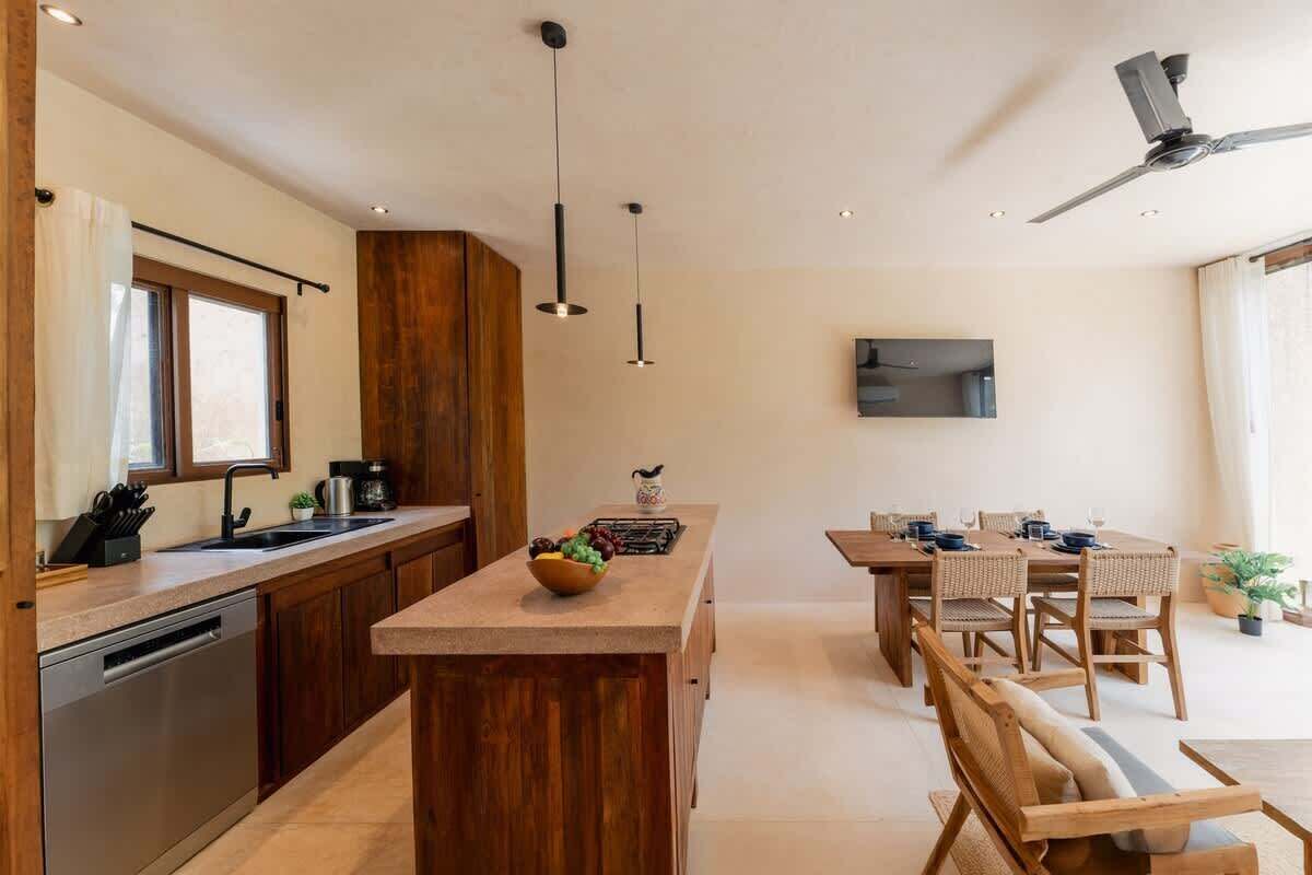 A modern kitchen with wooden cabinets, a central island, pendant lights, and a dining area with wooden furniture.