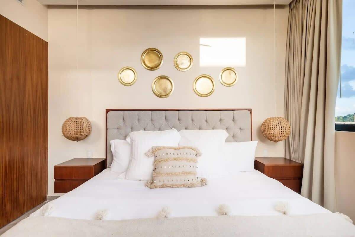 Um quarto com cabeceira estofada, cama com roupa de cama branca, duas mesas de cabeceira com luminárias trançadas e luminária fixada na parede.