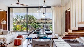 A modern dining room with a glass table, white upholstered chairs, and a view of greenery and buildings outside. - Luxury Penthouse with Private Rooftop Pool (Tulum)