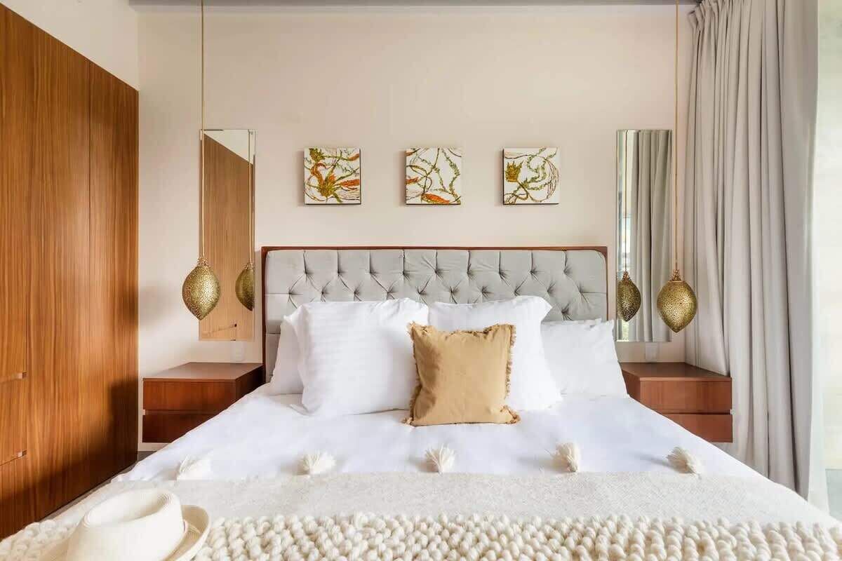 Quarto com cabeceira estofada, roupa de cama branca, almofada bege e três quadros abstratos na parede.