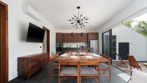 A modern dining area with a wooden table set for six, a TV mounted on the wall, and a kitchen in the background. - Luxury Jungle Villa with Private Pool & Concierge (Tulum)