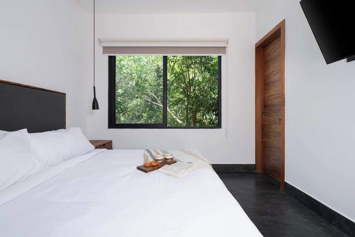 A modern bedroom with a large bed, a nightstand, a window with a view of trees, and a wooden door.