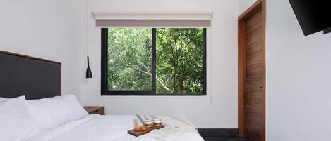 A modern bedroom with a large bed, a nightstand, a window with a view of trees, and a wooden door.