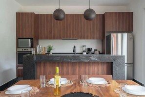 A modern kitchen with wooden cabinets, a granite countertop, and a dining area with a wooden table set for two. - Luxury Jungle Villa with Private Pool & Concierge (Tulum)
