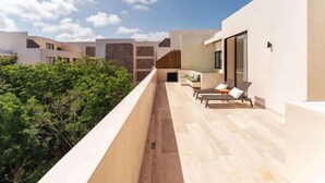 Ein moderner Balkon im Freien mit Blick auf üppiges Grün, einem Sitzbereich mit einer Couch und Lounge-Stühlen, und einem Gebäude mit einer Wand im Fischgrätenmuster.