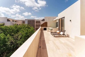 A modern outdoor balcony with a view of dense greenery, a seating area with a sofa and lounge chairs, and a building with a herringbone-patterned wall. - Sleek Penthouse Escape with Rooftop Pool (Tulum)