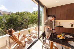 A modern kitchen with dark wood cabinets, a dining area with a table set for two, and a balcony with a view of trees. - Sleek Penthouse Escape with Rooftop Pool (Tulum)