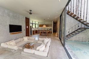 A spacious living area with a large sectional sofa, a flat-screen TV, and a kitchen in the background. - Esencia Villa - 3BR, Private Pool & Hammock (Tulum)