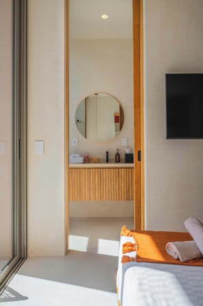 A hotel room with a bed, a wooden vanity with a mirror, a flat-screen TV mounted on the wall, and a window providing natural light. - Chic Aldea Zama Condo with Pool & Parking (Tulum)