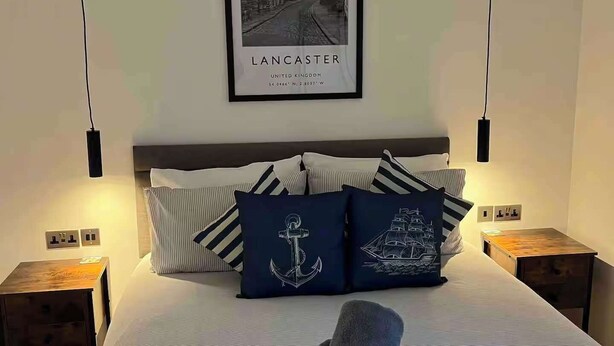 Cama bem arrumada com roupa de cama listrada, um quadro emoldurado de Lancaster e duas mesas de cabeceira com abajures.