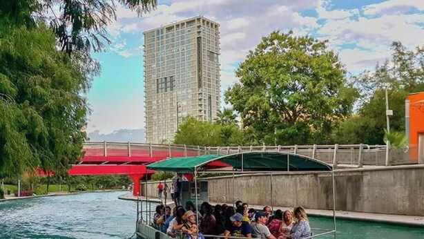 Un barco con pasajeros en un río, un puente y un edificio alto al fondo.