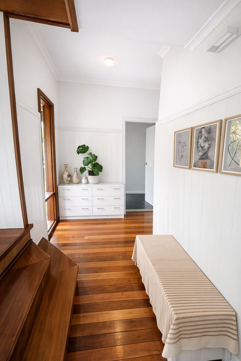 A hallway with wooden flooring, white walls, a bench, a plant, and framed artwork.