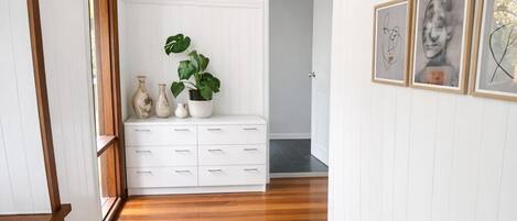 A hallway with wooden flooring, white walls, a bench, a plant, and framed artwork.
