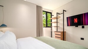 Ein modernes Schlafzimmer mit Bett, Wand-Fernseher, Fenster mit Blick auf Grünflächen und integriertem Kleiderschrank.
