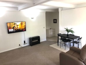 A living room with a flat-screen TV mounted on the wall, a black entertainment unit, a dining table with chairs, a potted plant, and a sofa.