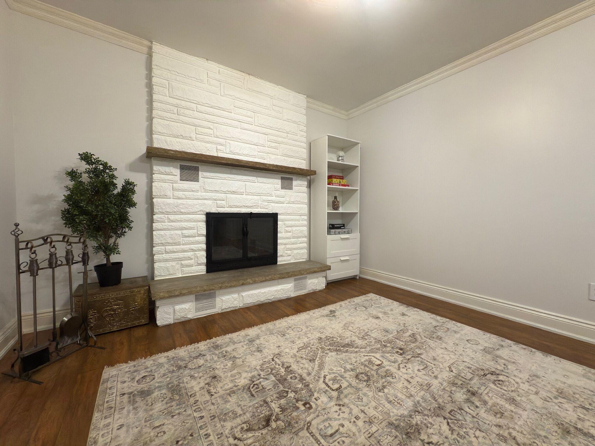 A living room with a white brick fireplace, a wooden bench, a potted plant, and a patterned rug.