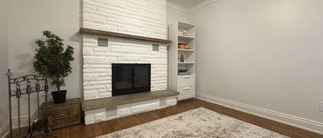A living room with a white brick fireplace, a wooden bench, a potted plant, and a patterned rug.