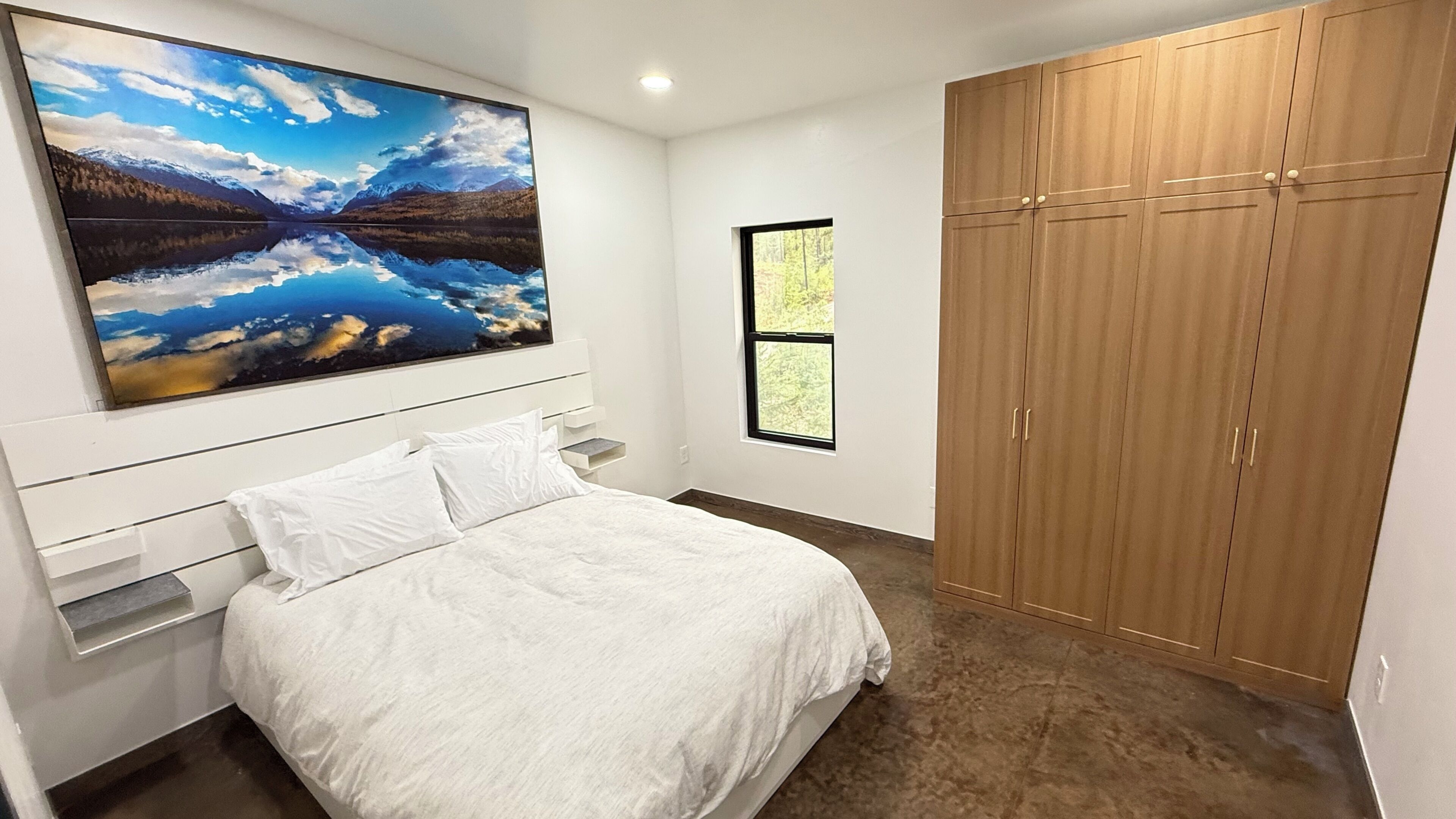 A bedroom with a bed, a large landscape painting, a wardrobe, and a window.