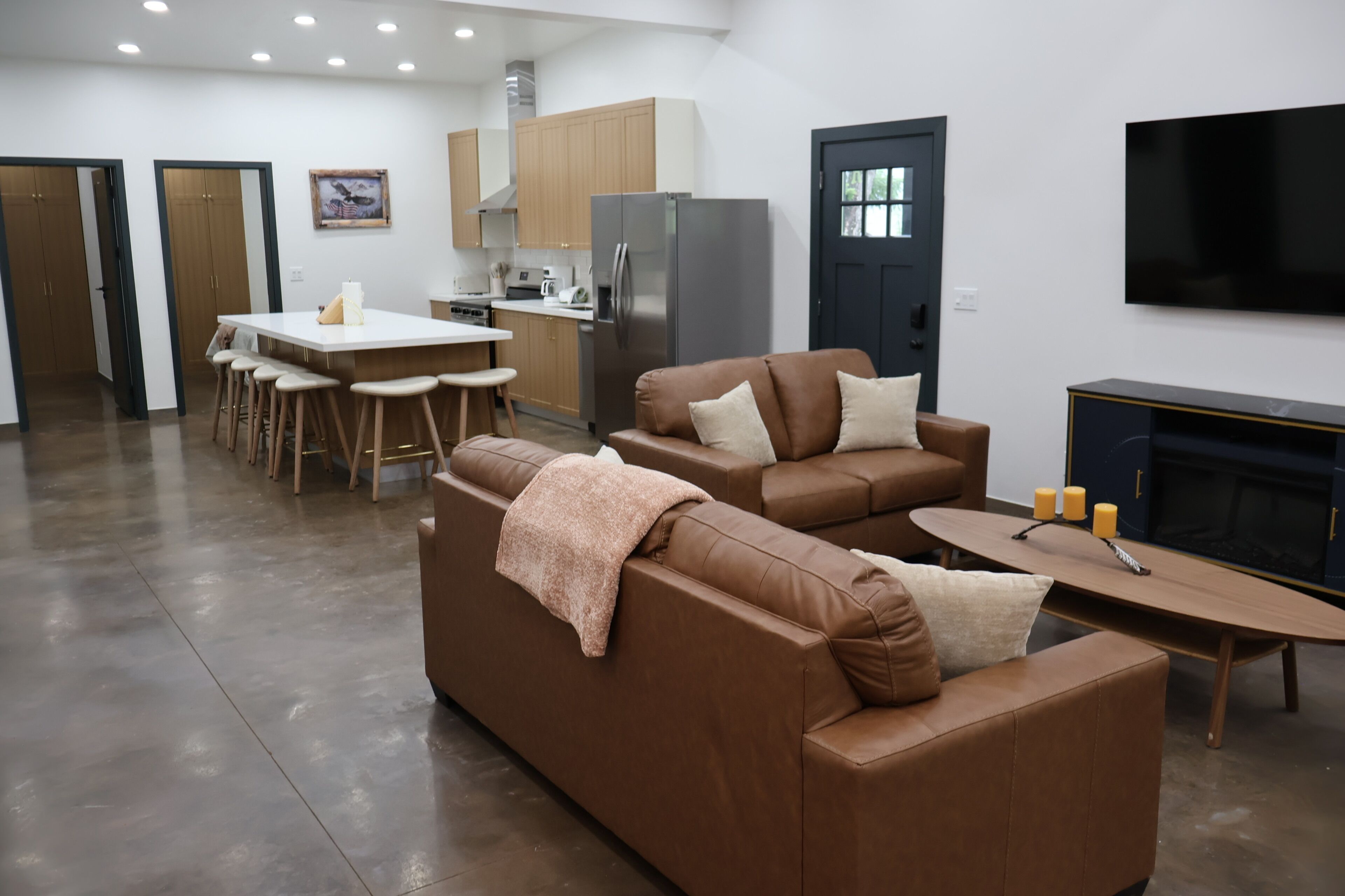A modern living room with a brown sofa, a dining area with stools, a kitchen with wooden cabinets, and a flat-screen TV mounted on the wall.