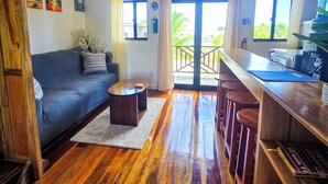 Sala de estar con sofá, mesa de centro y piso de madera. Cuenta con un balcón con barandilla y dispensador de agua.