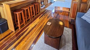 Una cocina moderna con armarios de madera, una cafetera y un televisor. Hay una zona de comedor con taburetes y una mesa redonda grande. El cuarto tiene piso de madera y un ventilador de techo.
