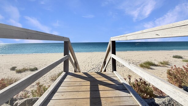Ein hölzerner Steg führt zu einem Strand unter blauem Himmel.