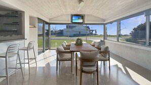 Ein moderner Essbereich mit einem Holztisch und -stühlen, einem an der Decke befestigten Fernseher und großen Fenstern mit Blick ins Freie.