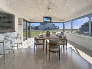 Ein moderner Essbereich mit einem Holztisch und -stühlen, einem an der Decke befestigten Fernseher und großen Fenstern mit Blick ins Freie.