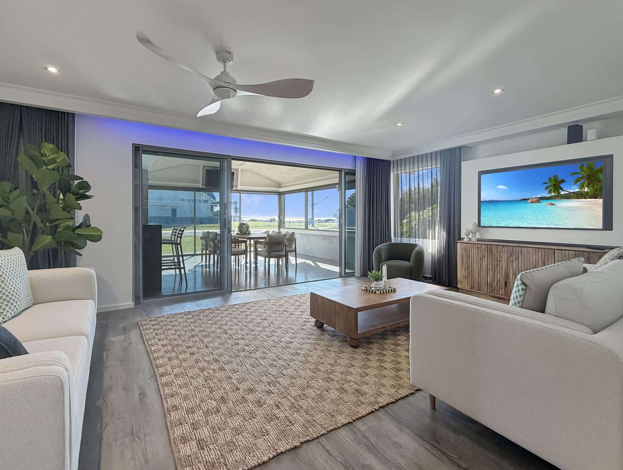 Uma sala de estar moderna com ventilador de teto, TV de tela plana, área de jantar com mesa e cadeiras, e um grande painel de vidro deslizante que dá para um espaço ao ar livre.