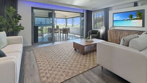 Ein modernes Wohnzimmer mit Deckenventilator, Flachbildfernseher, Essbereich mit Tisch und Stühlen sowie eine große Schiebeglastür, die zu einem Außenbereich führt.