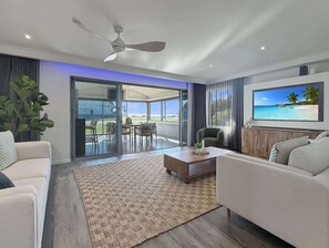 Ein modernes Wohnzimmer mit Deckenventilator, Flachbildfernseher, Essbereich mit Tisch und Stühlen sowie eine große Schiebeglastür, die zu einem Außenbereich führt.