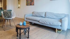 Ein Wohnzimmer mit grauem Sofa, einem runden Holztisch mit blauen Vasen, einem modernen Sessel mit gemustertem Kissen und einem Spiegel an der Wand.