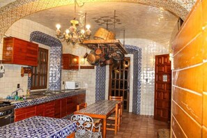 Une cuisine avec des carreaux décoratifs, des armoires en bois, un porte-ustensiles suspendu et un coin repas avec une nappe à motifs.