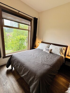 Ein Schlafzimmer mit großem Fenster, einem Bett mit grauer Decke, einem Nachttisch mit Lampe und Blick ins Grüne.