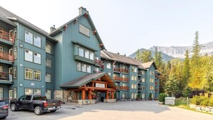 A multi-story building with a prominent entrance, balconies, and a mountainous backdrop. - Spectacular Ski-in/Ski-out Mountain Condo with A/C! (Fernie)