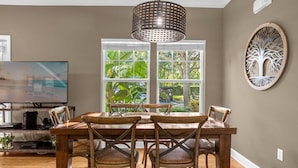 Uma área de refeições com uma mesa e cadeiras de madeira, um lustre, uma televisão e uma janela com vista para o verde.