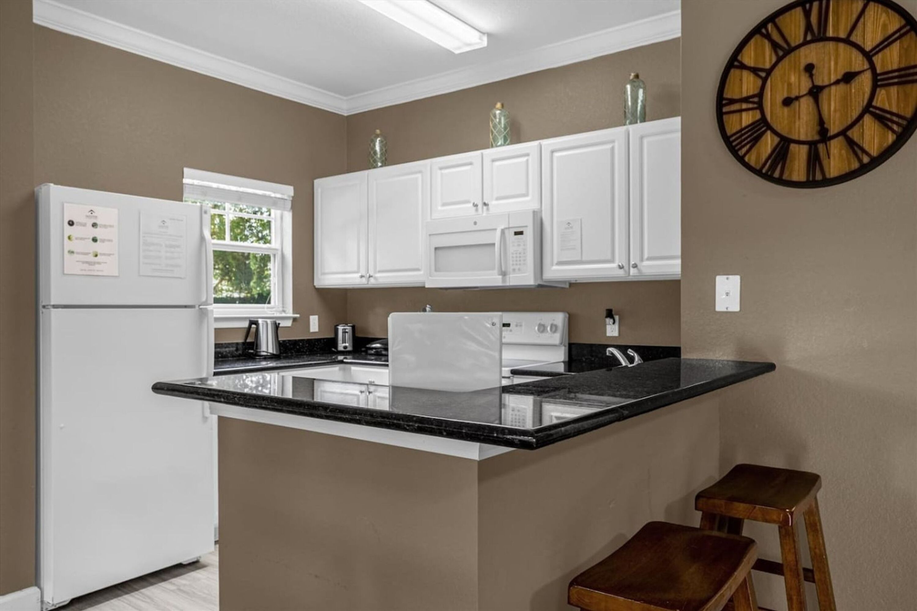Una cocina con armarios blancos, microondas, refrigerador, reloj y ventana con follaje en el exterior.