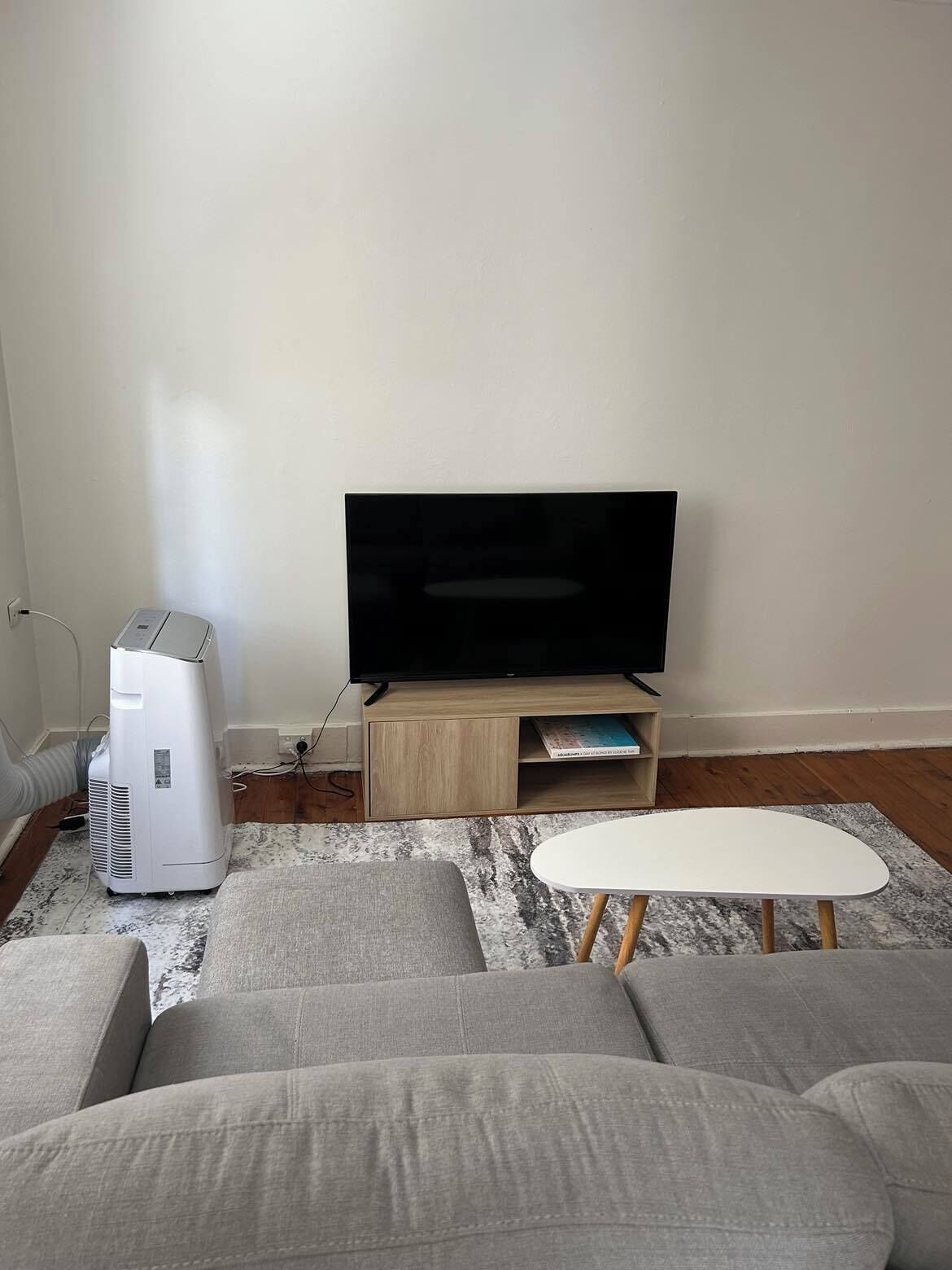 Ett vardagsrum med en platt-TV, en vit luftkonditioneringsanläggning, ett TV-bord i trä, en grå soffa och ett litet vitt bord.