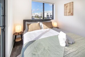 Ein Hotelzimmer mit einem Bett, zwei Nachttischen mit Lampen, einem Fenster mit Blick auf Gebäude und einem Wandbild.
