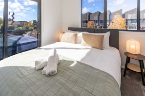 Ein Hotelzimmer mit einem großen Bett, einem Nachttisch mit Lampe und Blick auf Gebäude durch das Fenster.