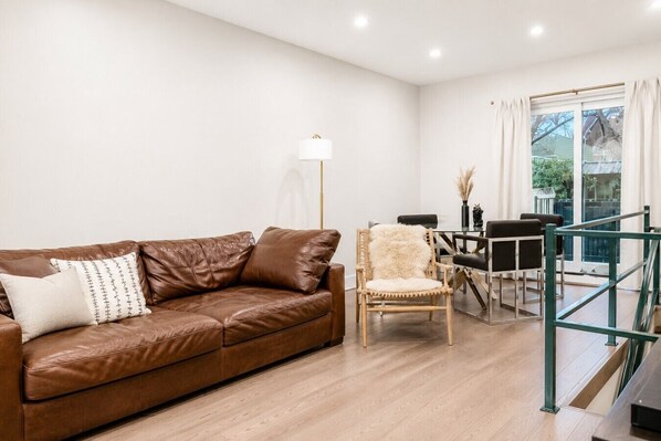 A living room with a brown leather sofa, a white fluffy chair, a glass table with chairs, and a floor lamp. - Stunning 3BD Duplex with Private Garden Oasis (New York)