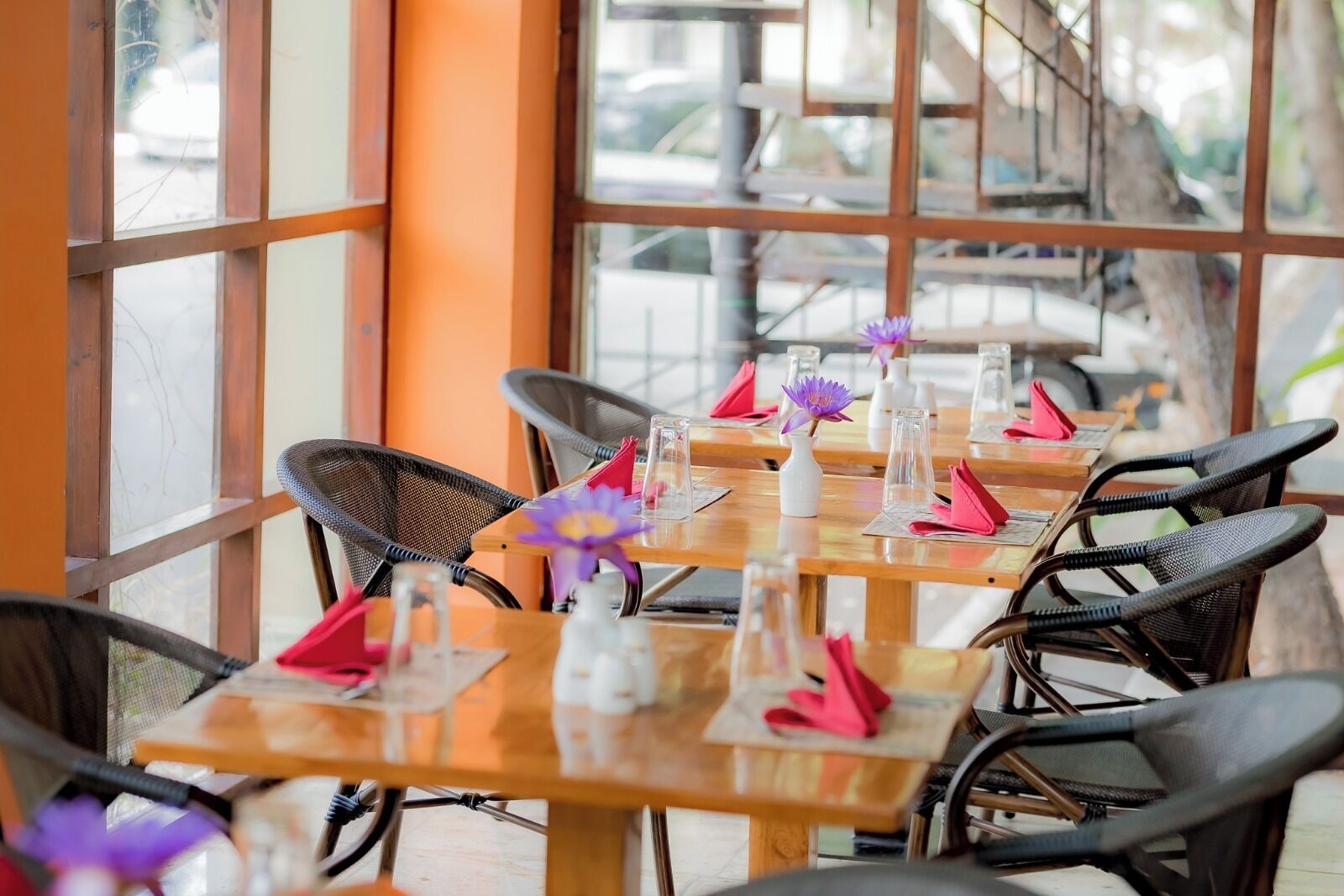 A row of wooden tables with chairs, each table set with a clear glass, a pink napkin, and a purple flower.